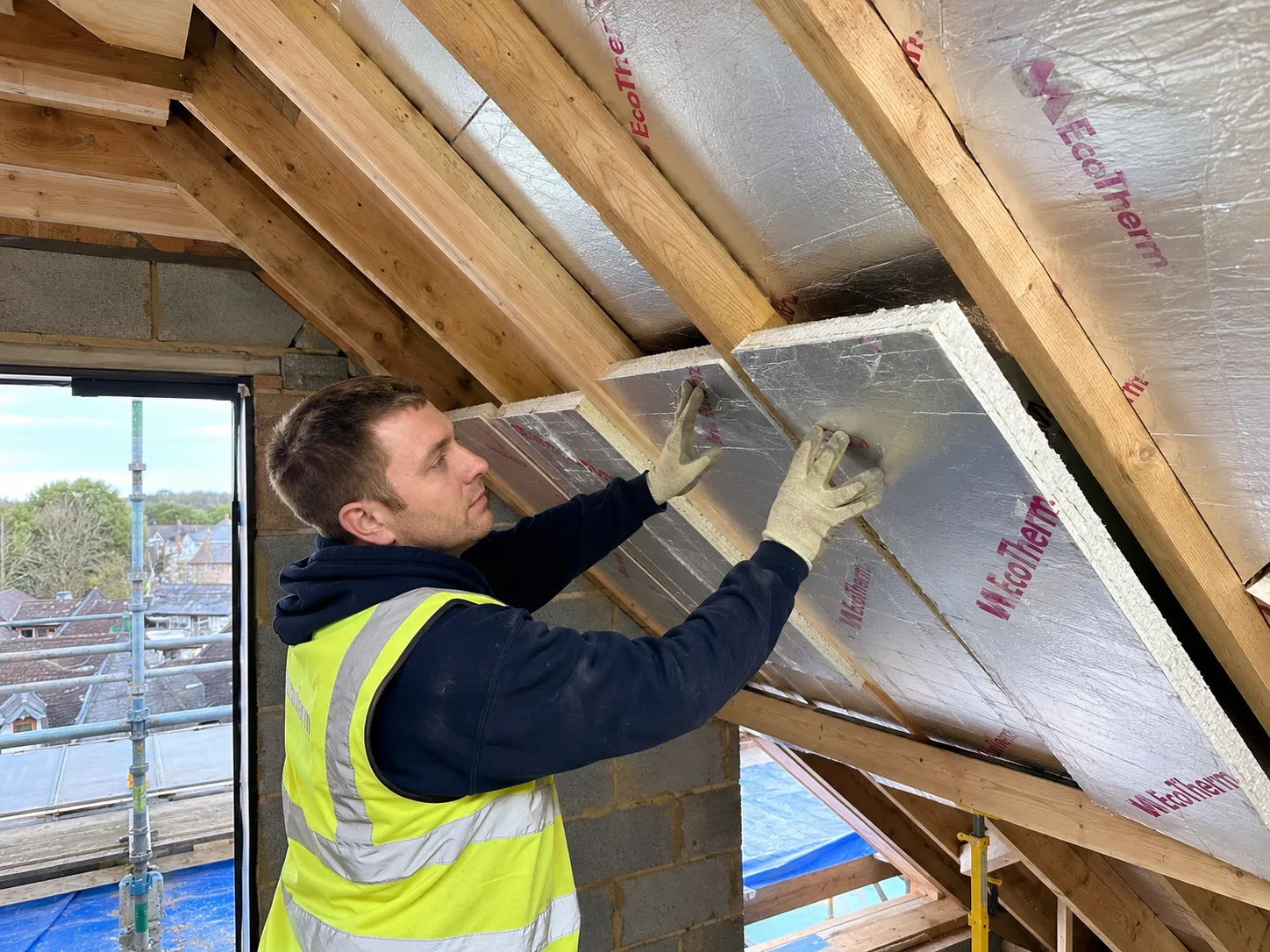 Professional builders installing foil-backed rigid PIR insulation between loft rafters to meet regulations.